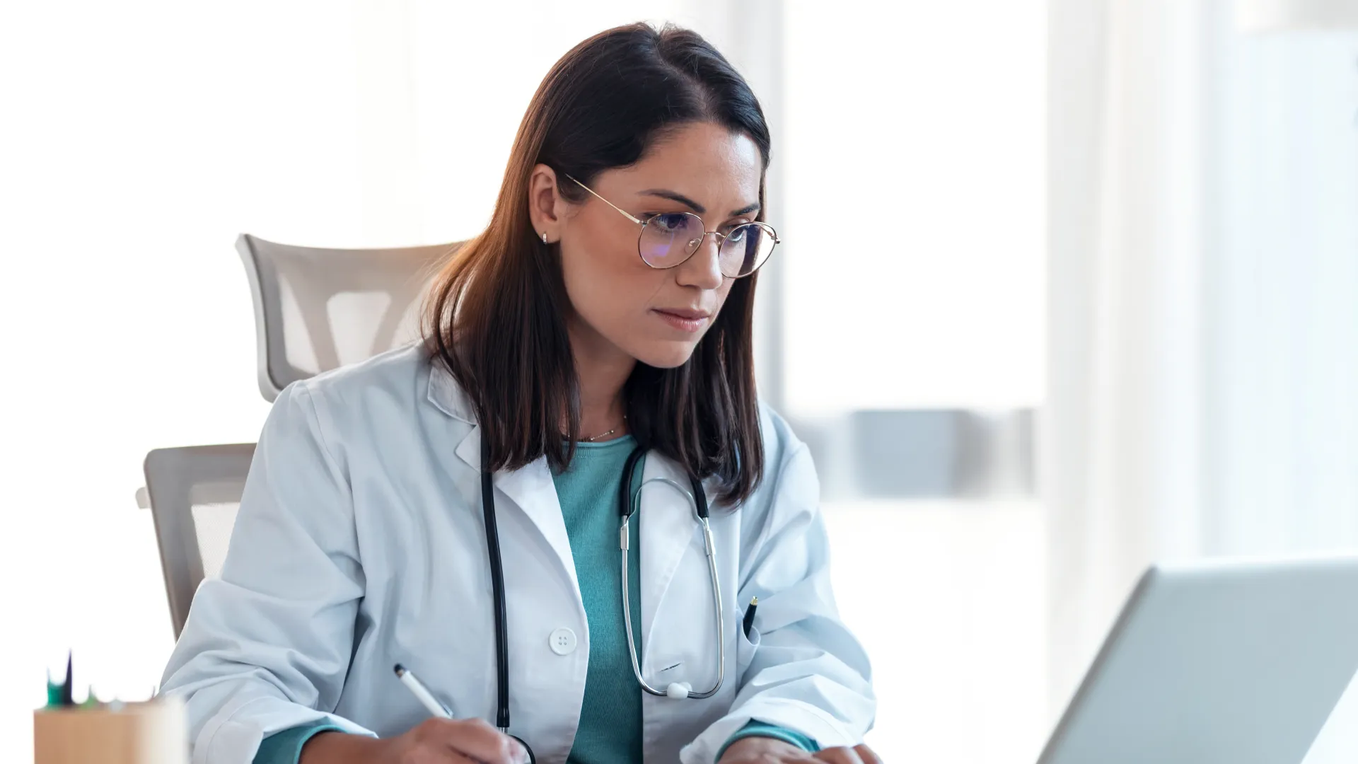 Female doctor in white coat works on laptop, taking notes in modern office setting.