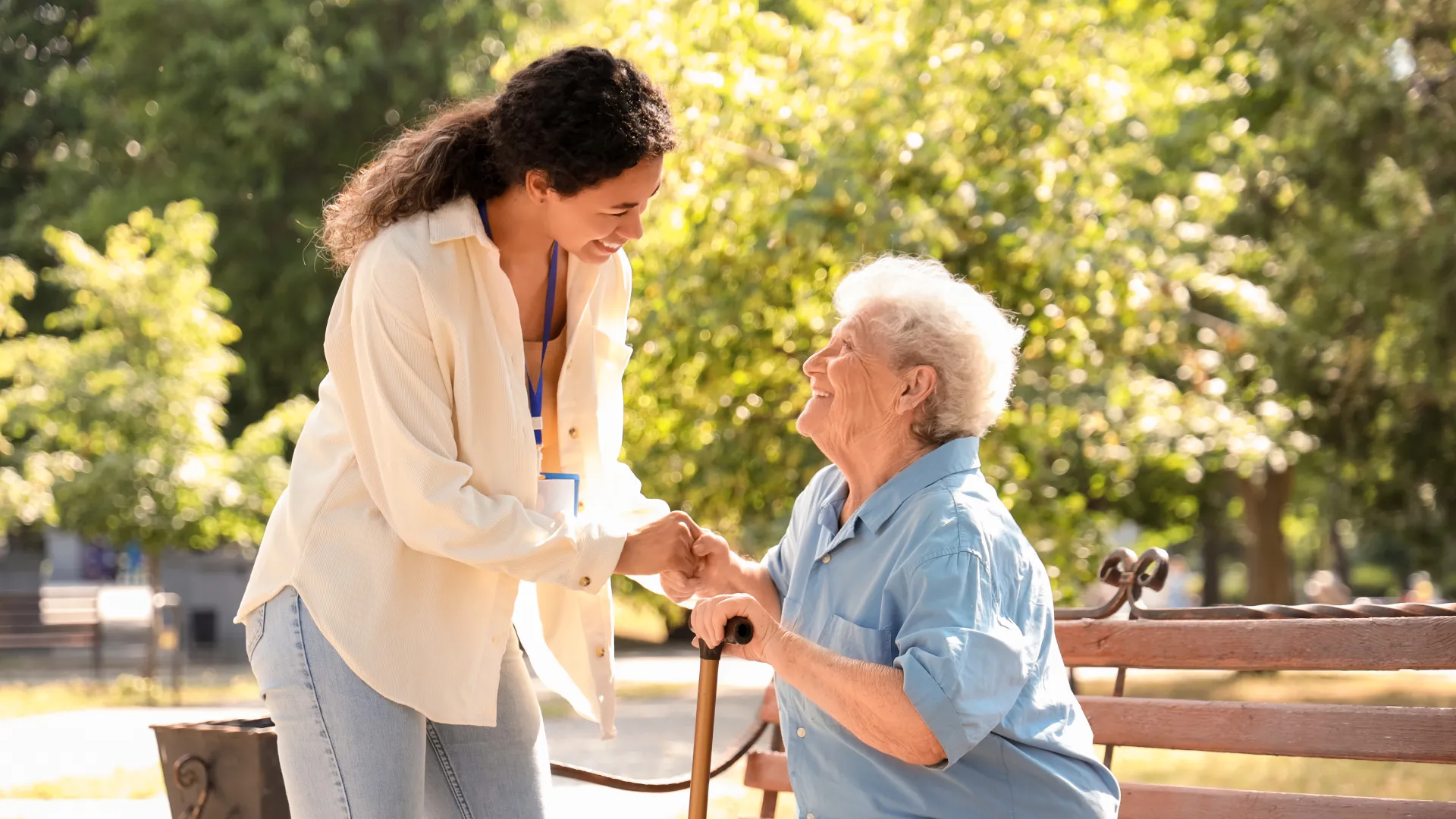 Caregiver assisting elderly woman on a park bench, surrounded by trees in a warm, sunny setting.