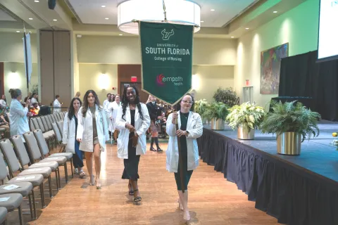 Nursing students in white coats march holding University of South Florida College of Nursing banner at ceremony.