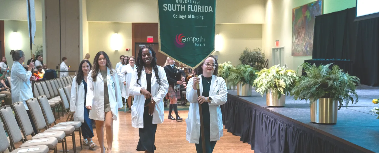Nursing students in white coats march holding University of South Florida College of Nursing banner at ceremony.