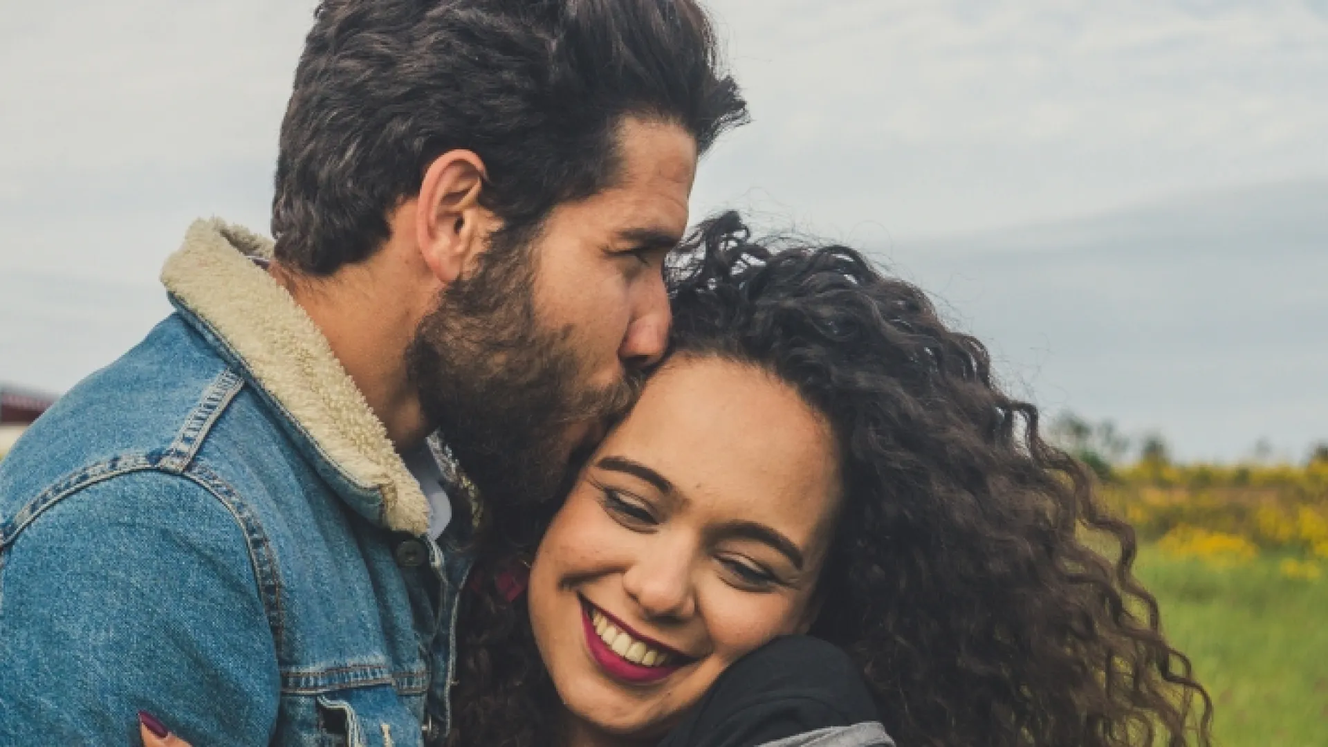 Couple embracing outdoors with man kissing woman on forehead in a green field under cloudy sky