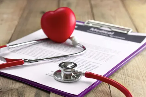 Red stethoscope with a heart-shaped object resting on medical documents clipped to a purple clipboard.