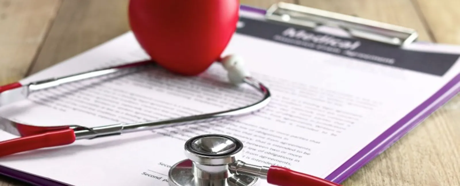 Red stethoscope with a heart-shaped object resting on medical documents clipped to a purple clipboard.