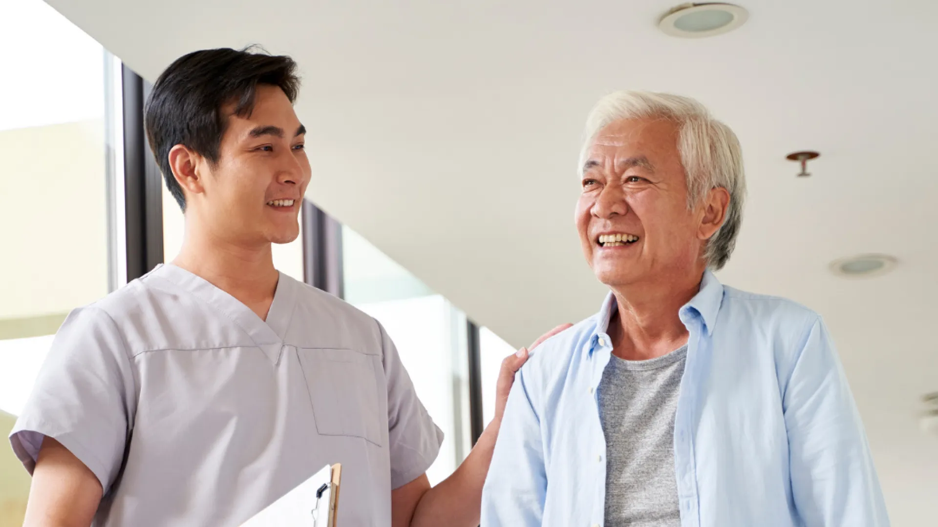 Healthcare professional assisting elderly man with walker during physical therapy session in bright clinic hallway
