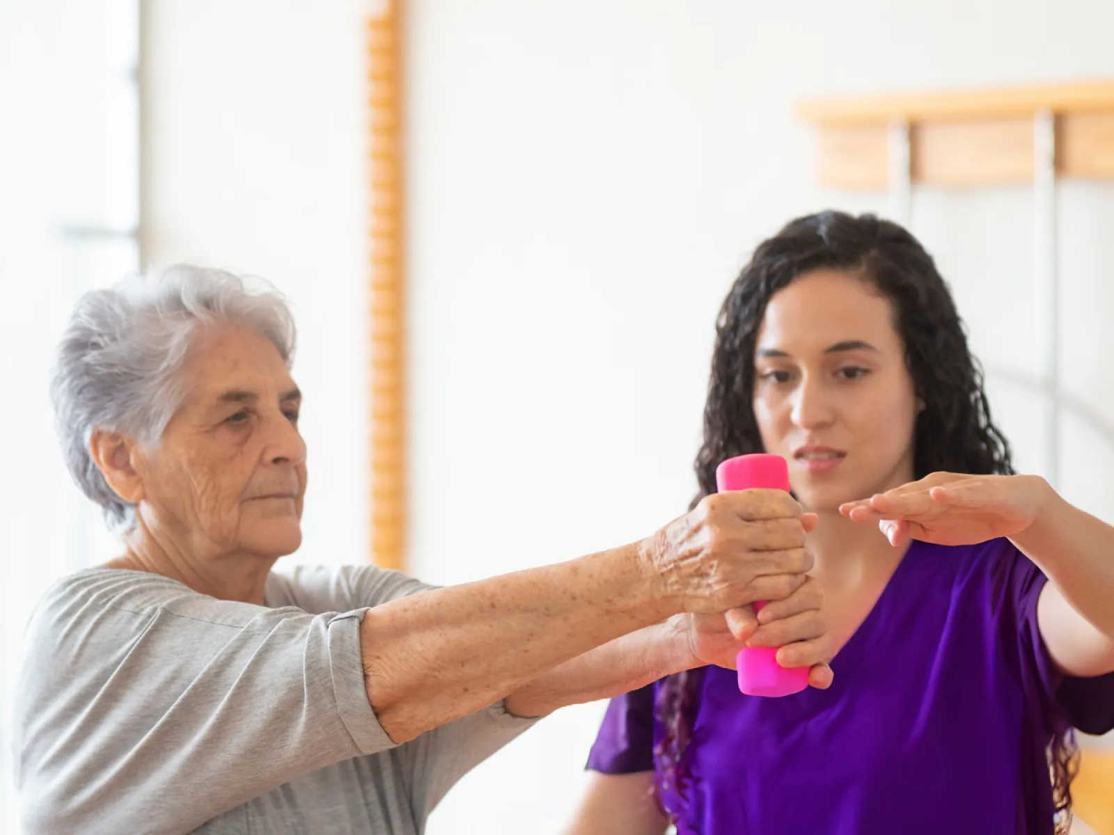 Senior woman doing arm exercises with a pink dumbbell guided by a physical therapist in purple shirt.