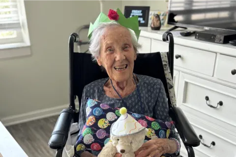 Elderly woman in wheelchair smiling, wearing a paper crown, holding a birthday-themed pillow and teddy bear in living room.