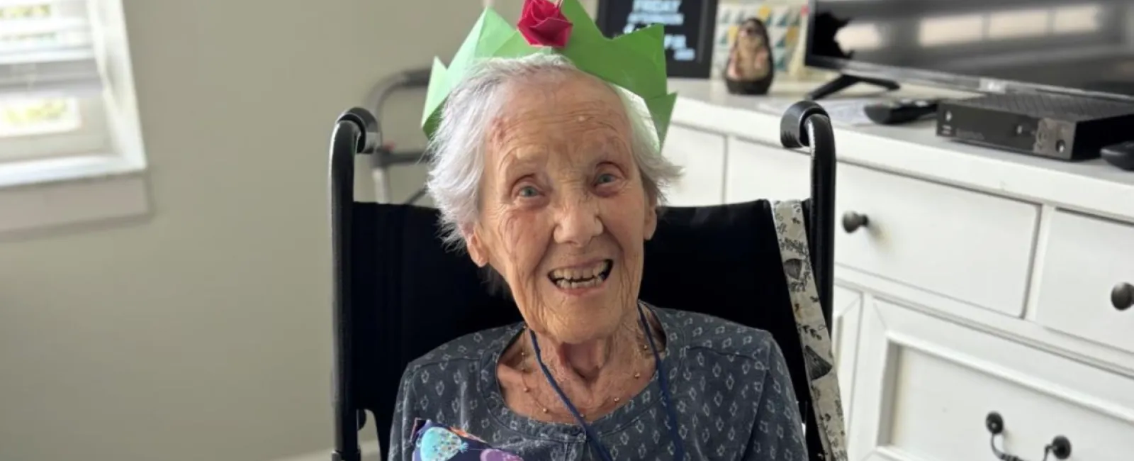 Elderly woman in wheelchair smiling, wearing a paper crown, holding a birthday-themed pillow and teddy bear in living room.