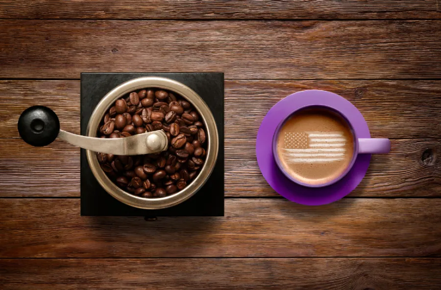 Top view of a coffee grinder with beans and a purple cup of coffee with American flag foam art on wooden table
