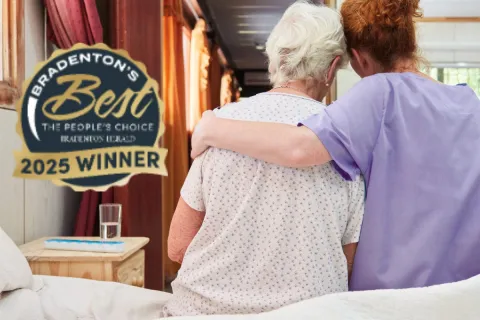 Caregiver in purple scrubs embraces elderly woman in bedroom setting, showing support and comfort.